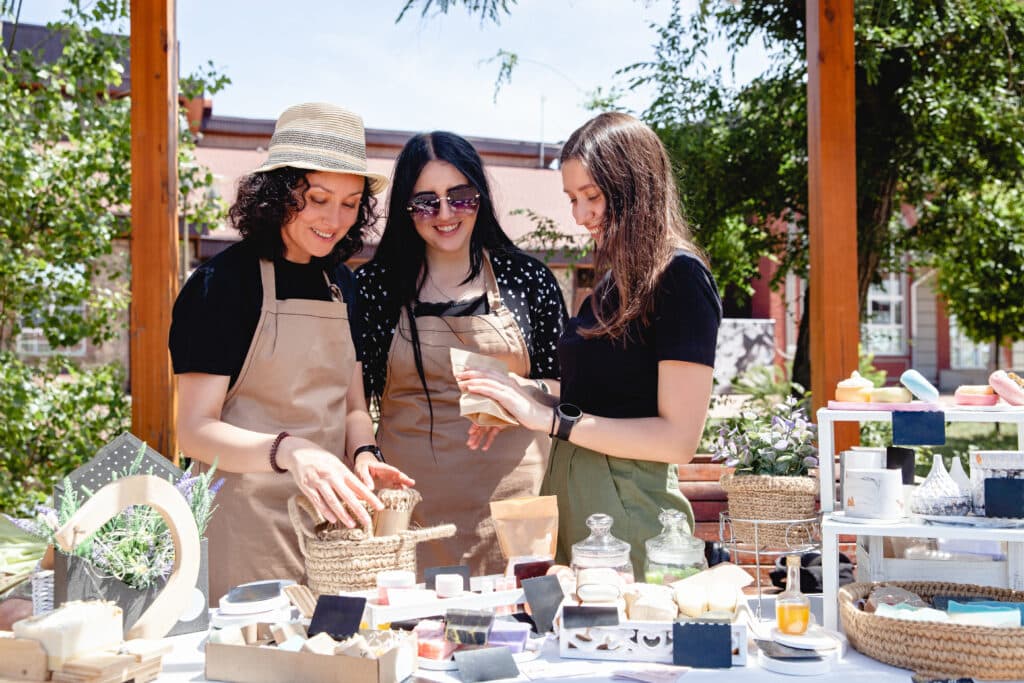 Three brunette women sellers of European appearance in aprons at a street fair laughing discuss their goods.